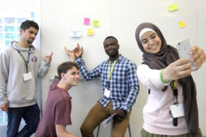 Student takes a selfie with three other students in front of a whiteboard with post-it notes