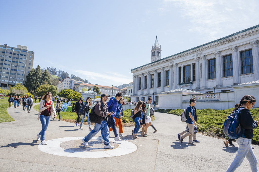 Students walk across the Berkeley campus with a academic building and the Campanile in the background.