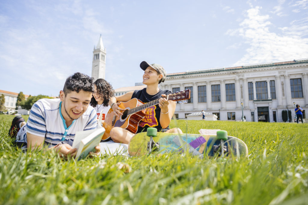 student plays guitar on grass next to two other students