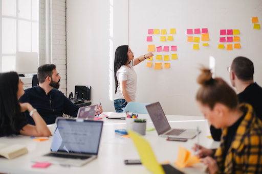 Alt text: Four students sit at a conference table with laptops. They are all looking at a fifth person who is sticking post-its on a whiteboard.