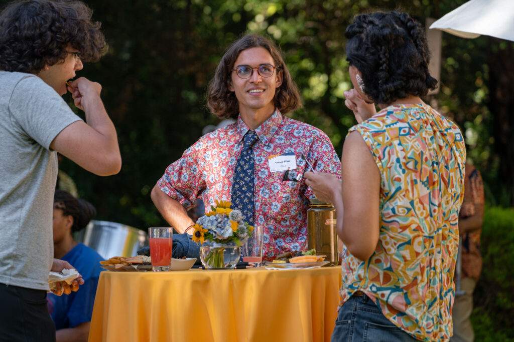 A grad student stands at a table speaking with other students.