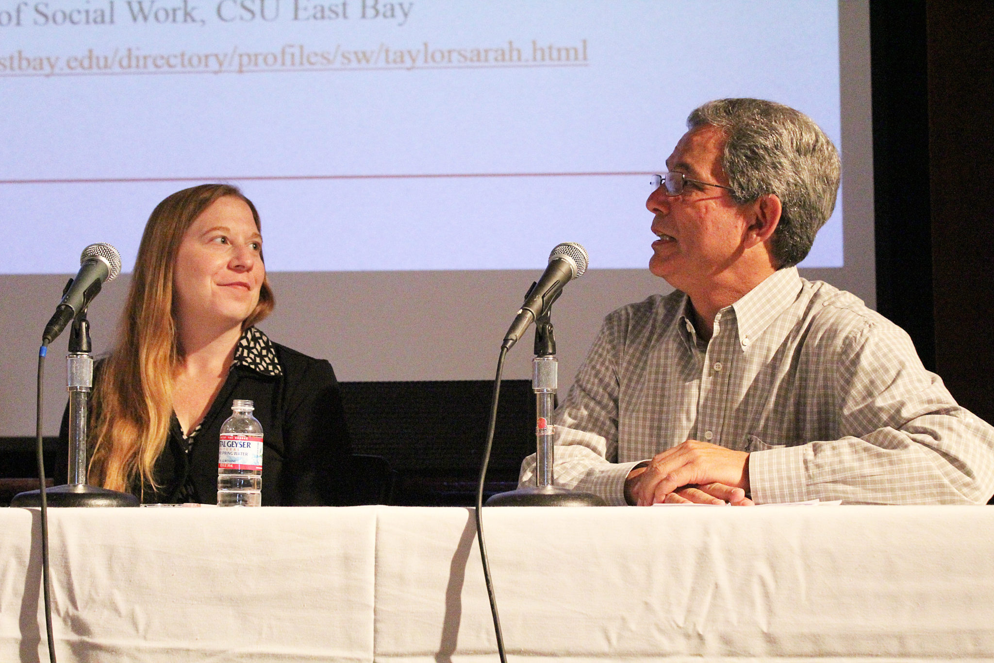 Photo of two people sitting at a table in front of a microphone.