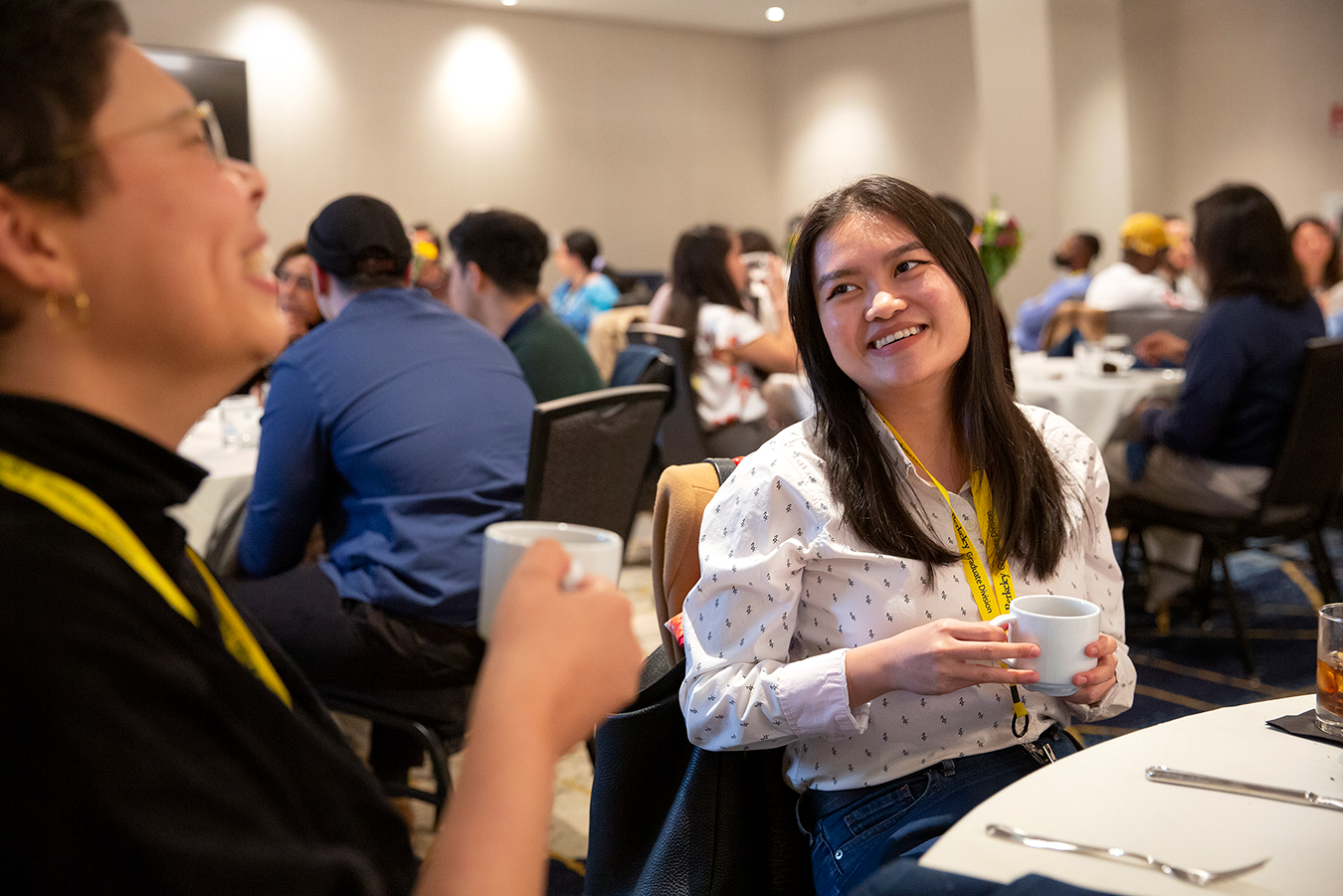 female student in white shirt smiling at another student in blue shirt while sitting at a table during a conference